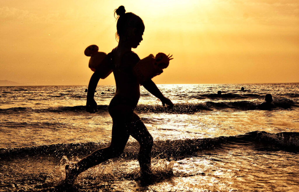 Kind mit Schwimmflügeln läuft am Ufer des Meeres bei Sonnenuntergang, Silhouetten von Menschen im Wasser im Hintergrund