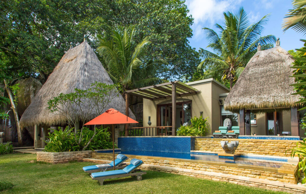 Tropische Resort mit Strohdächern, Pool mit blauen Kacheln, Terrasse mit Liegestühlen und Sonnenschirm auf den