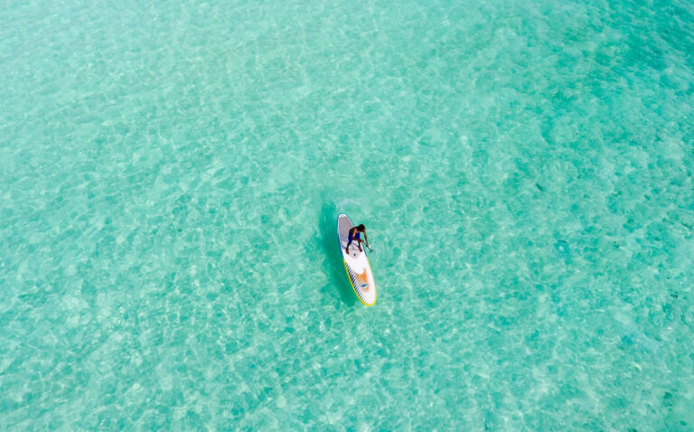 Person auf weißem Stand-Up-Paddleboard mit gelben Akzenten auf ruhigem, klarem türkisfarbenem Meer, von oben fotografiert
