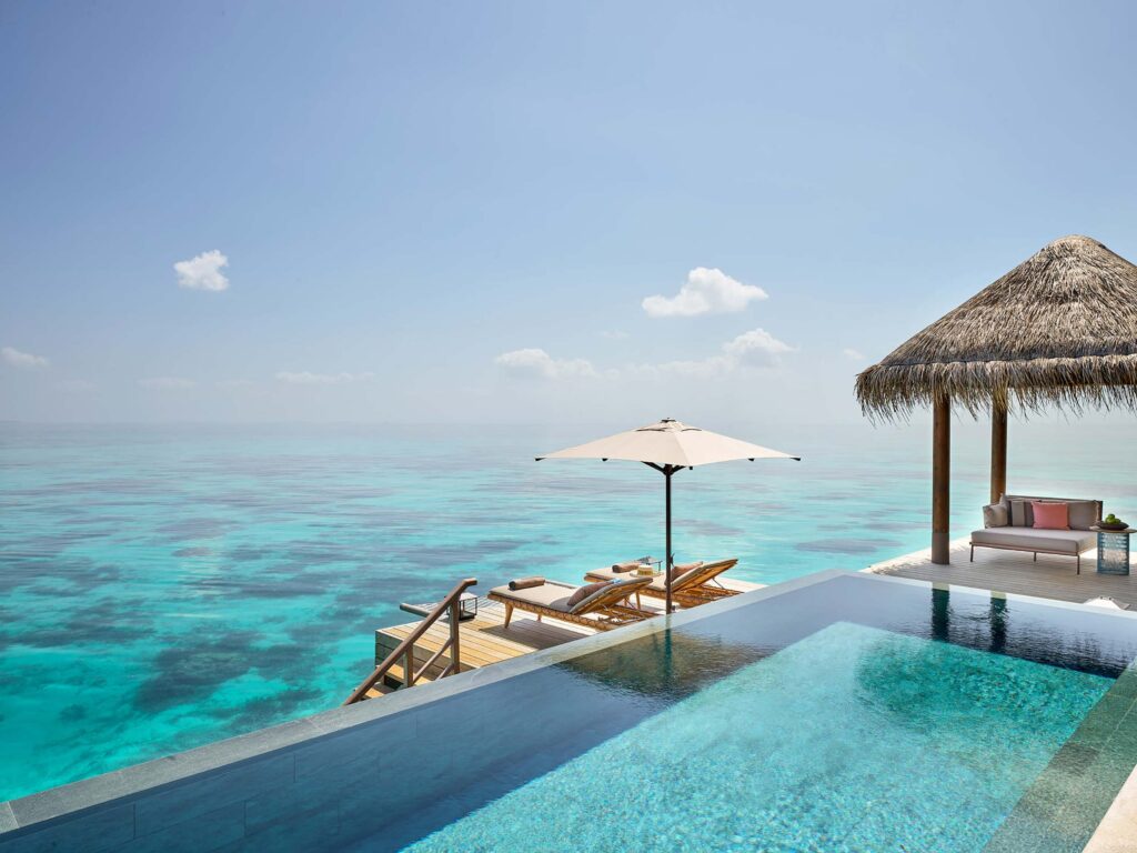 Infinity-Pool mit türkisfarbenem Meer im Hintergrund, Terrasse mit Strohdachpavillon, Sofa, Liegestühlen und Sonnenschirm
