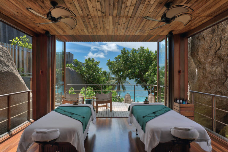 Spa-Raum mit zwei Behandlungsliegen, Holzboden und Glaswänden mit Blick auf tropischen Garten und Meer