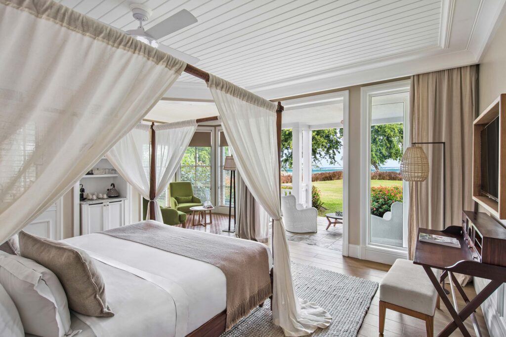 Hotelzimmer mit weiß drapiertem Himmelbett, grünem Sessel, Holzboden und Blick auf Garten und Meer in Mauritius