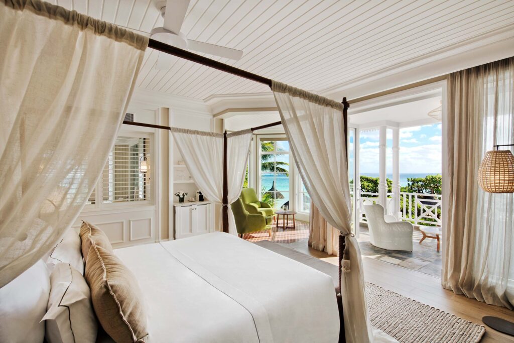 Hotelzimmer mit Himmelbett, cremefarbenen Vorhängen, Holzboden, Balkon mit Meerblick und weißen Sitzmöbeln