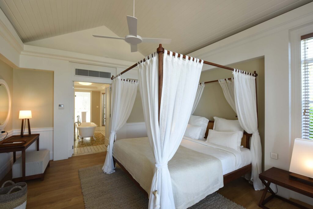 Geräumiges Hotelzimmer mit Himmelbett, weißen Vorhängen, Holzmöbeln und freistehender Badewanne im Hintergrund