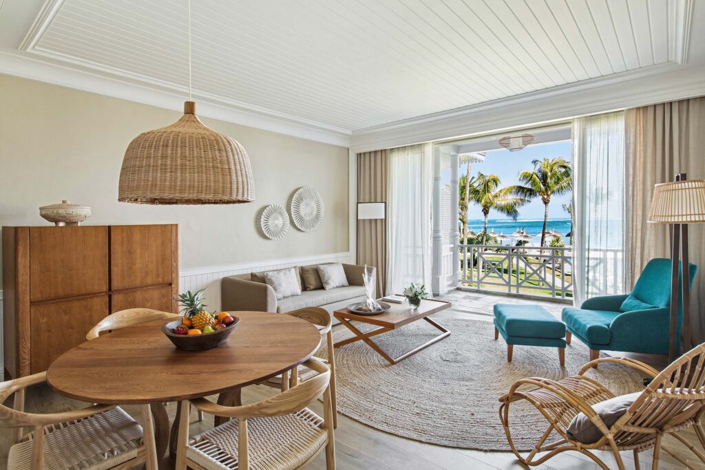 Hotelzimmer mit ovalem Holztisch, geflochtenen Stühlen, türkisfarbenem Sofa und Blick auf Meer und Palmen