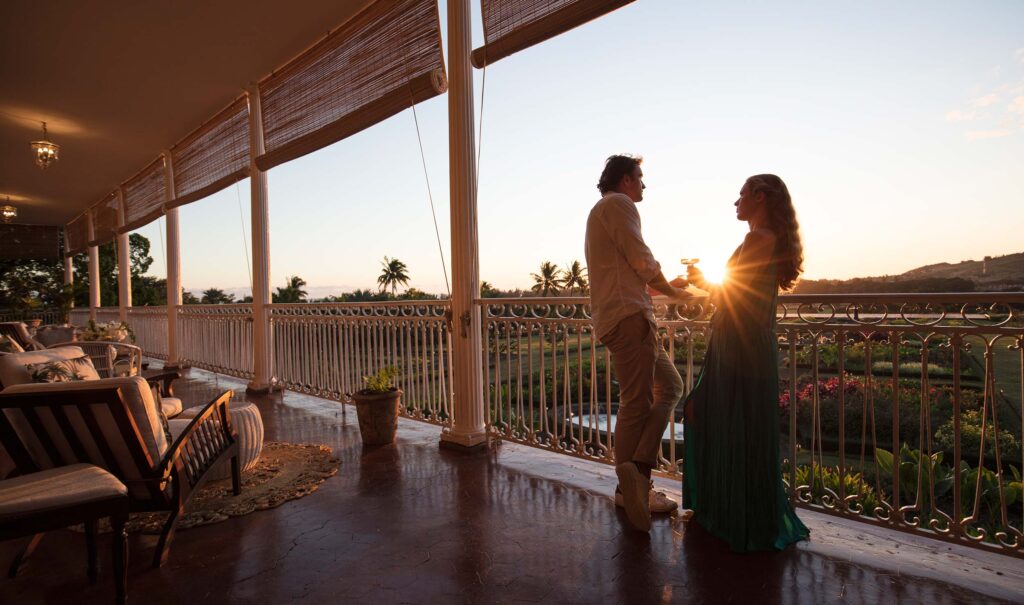Veranda mit weißen Eisenbalustraden, Holzsesseln und Polstermöbeln, Blick auf tropische Gärten und Sonnenuntergang