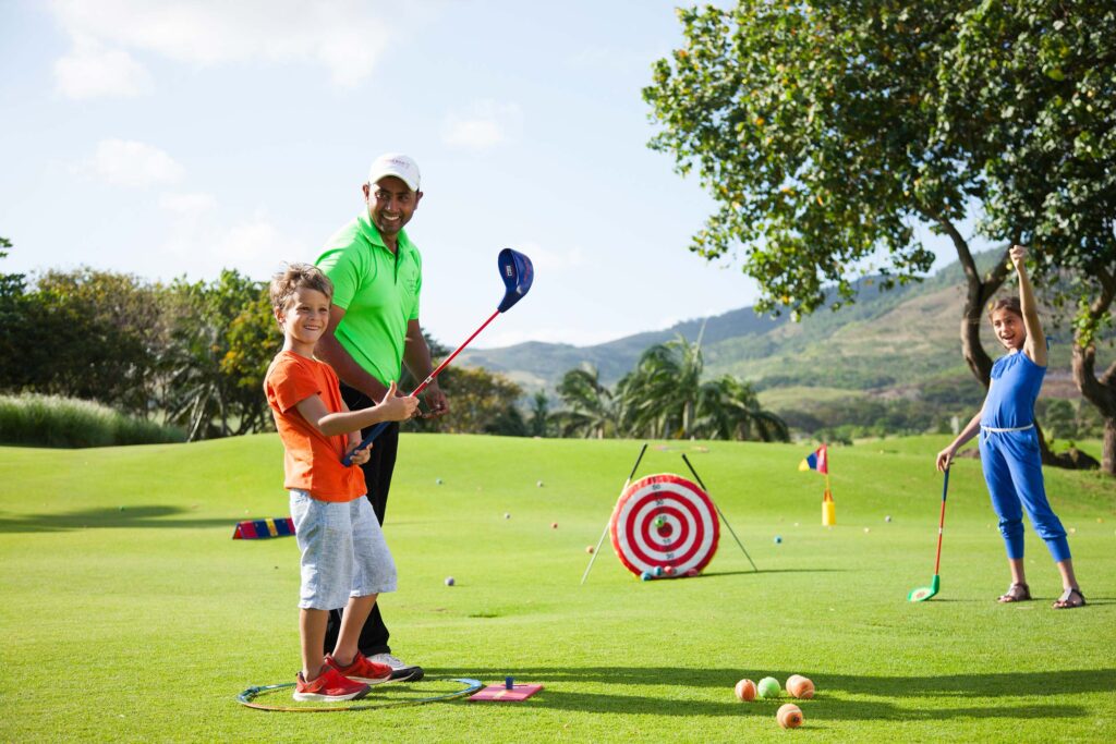 Mann und zwei Kinder mit Golfschlägern auf grüner Golflandschaft vor Bäumen und Hügeln bei Heritage Le Telfair