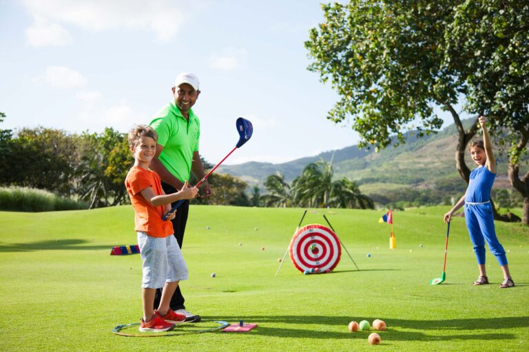 Mann und zwei Kinder mit Golfschlägern auf grüner Golflandschaft vor Bäumen und Hügeln bei Heritage Le Telfair