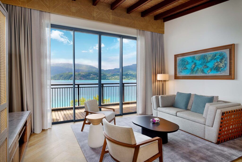 Hotelzimmer mit Sofa, zwei Stühlen, Couchtisch, großem Fenster und Blick auf Wasser und bewaldete Hügel
