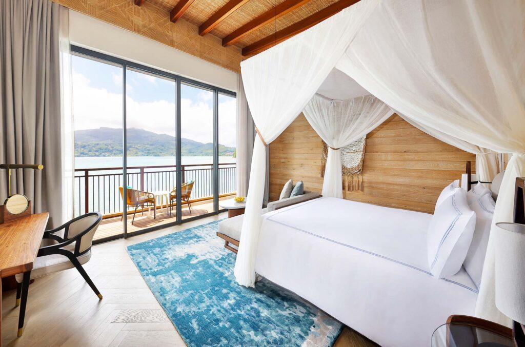 Hell eingerichtetes Hotelzimmer mit Himmelbett, weißen Vorhängen, Holzverkleidung und Blick auf Balkon mit See und Wald