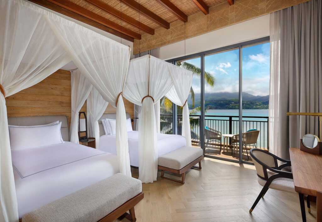 Hotelzimmer mit zwei Himmelbetten, Holzelementen und Balkon mit Blick auf Bucht und Palmen