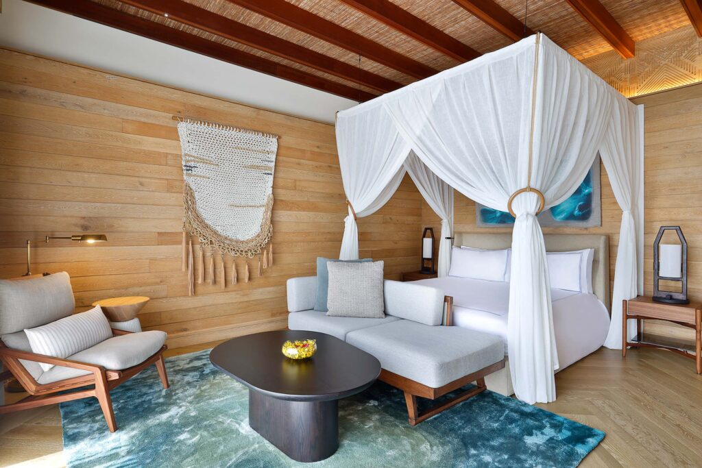 Hotelzimmer mit Himmelbett, weißen Vorhängen, Holzwänden, blau-grünem Teppich, grauem Sofa und Makramee-Dekoration