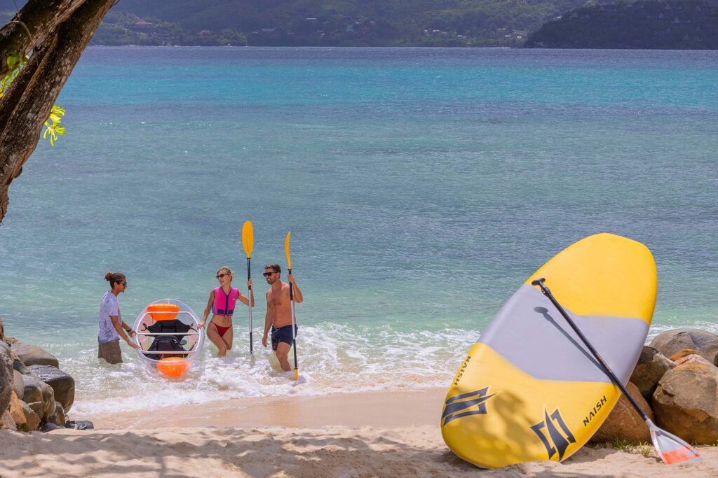 Drei Personen gehen mit halb-transparentem Kajak am Strand an Land, gelb-graues Paddleboard lehnt an Felsen, Meer.