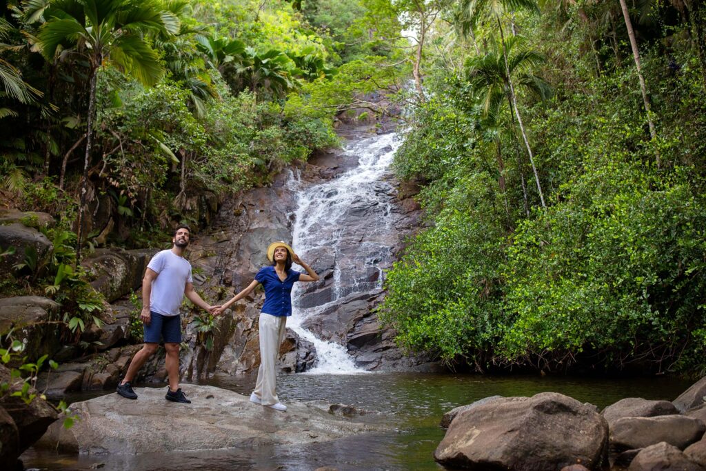 Wasserfall über Felsen mit zwei Personen auf großem Stein, umgeben von grünen Bäumen