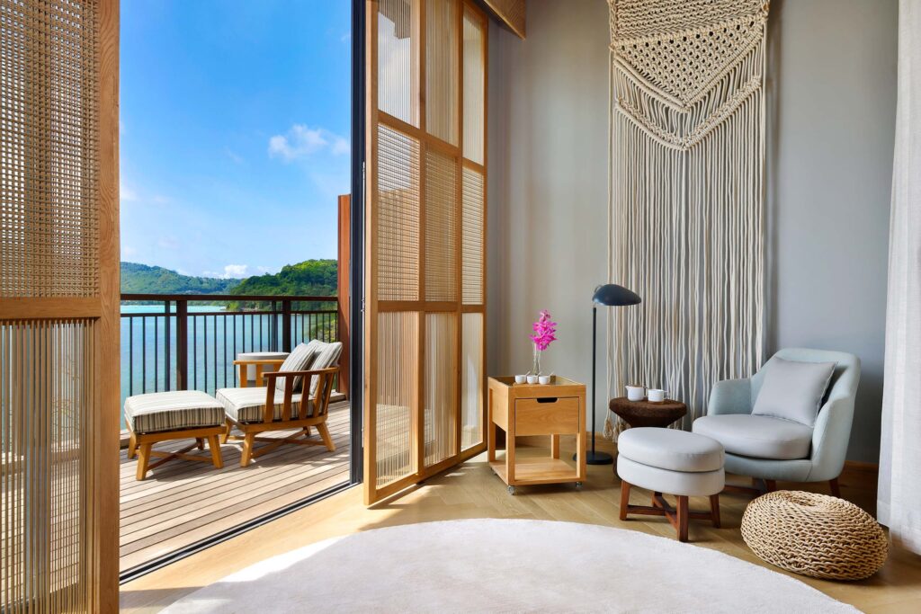 Hotelzimmer mit offenem Balkon, Blick auf Wasserlandschaft und bewaldete Hügel, Holzmöbel und Makramee-Dekor