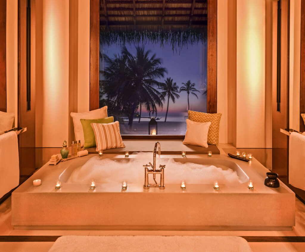 Badezimmer mit großer eingelassener Badewanne, Kerzen, Kissen und Fenster mit Blick auf Palmen bei Dämmerung