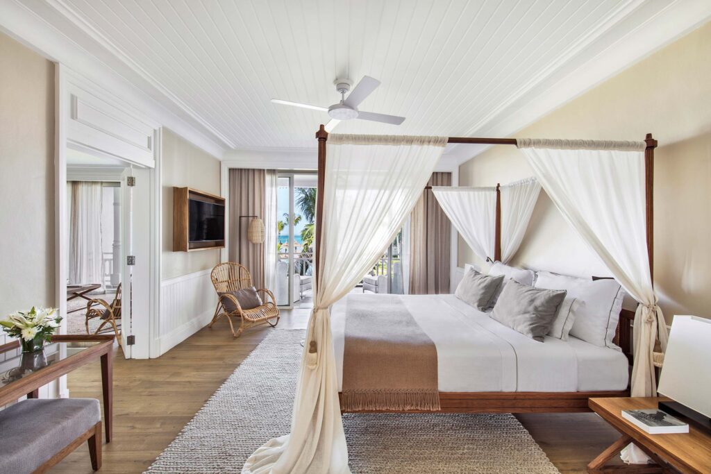 Hotelzimmer mit großem Himmelbett, weißen Vorhängen, Rattanstuhl, Holztisch und Blick auf Palmen in Mauritius