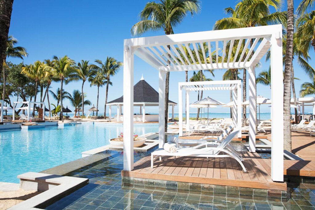 Hotelpool mit klarem blauem Wasser, weißen Holzpavillons, Liegestühlen und hohen Palmen im Heritage Le Telfair auf Mauritius