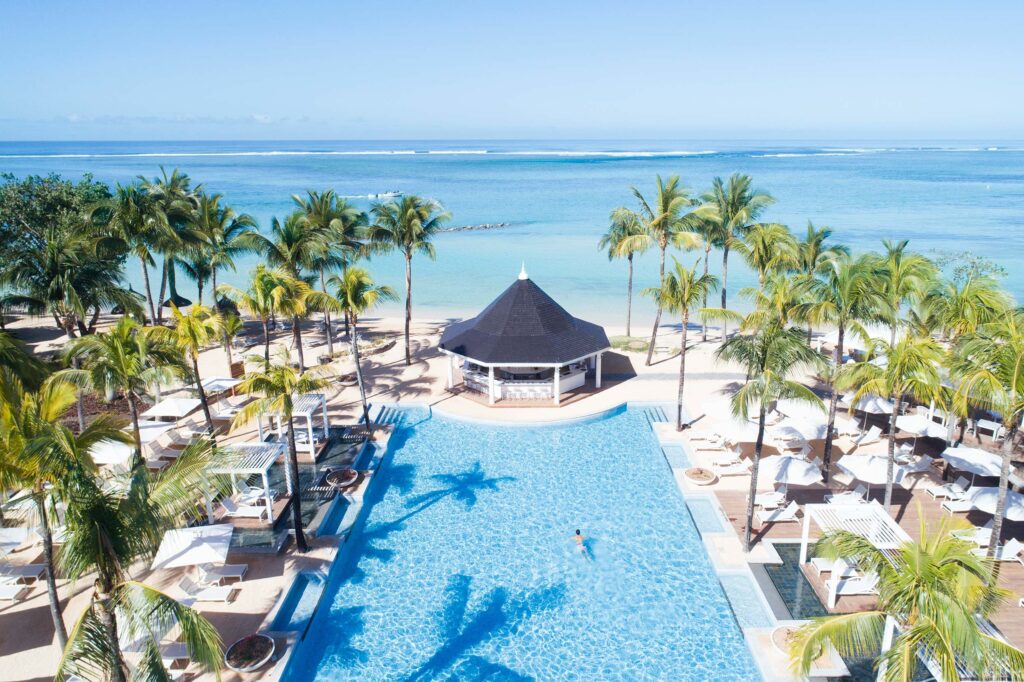 Großer rechteckiger Pool mit blauem Wasser, Palmen, weißem Sonnenschirmen und schwarzem Pavillon am Strand
