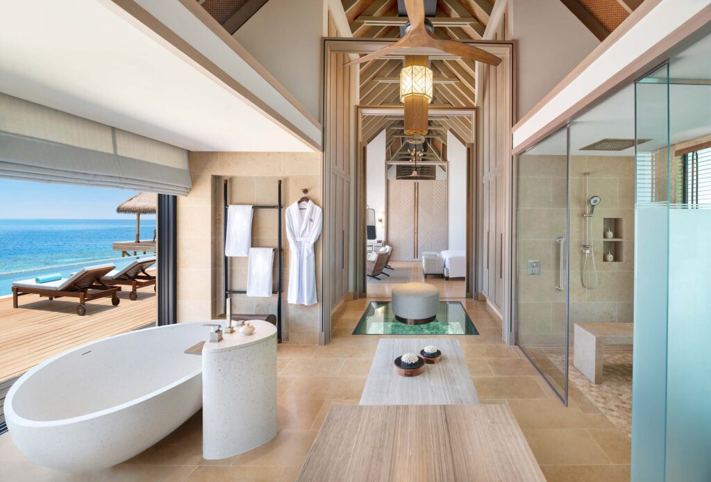 Hotelzimmer mit moderner Badewanne aus hellem Stein, gläserner Dusche, großen Fenstern und Terrasse mit Meerblick