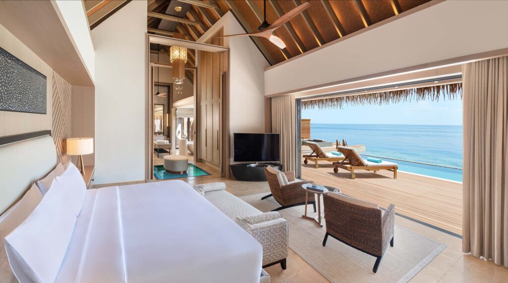 Hotelzimmer mit großem Bett, zwei geflochtenen Sesseln, Glasfront und Blick auf Terrasse mit Pool und Meer