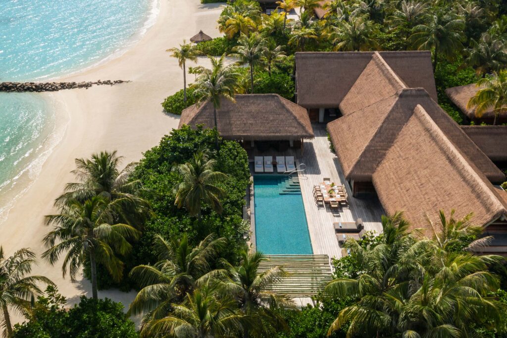 Vogelperspektive auf Resort mit strohgedeckten Gebäuden, Pool, Liegestühlen und Palmen am Strand