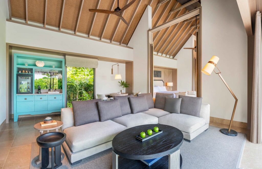 Modernes Hotelzimmer mit hellgrauem Sofa, rundem Holztisch, türkisfarbener Minibar und Blick auf tropisches Grün