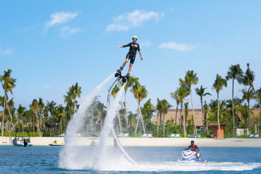 Person auf Flyboard und weitere auf Jetski vor Palmen und weißem Sandstrand bei Tageslicht