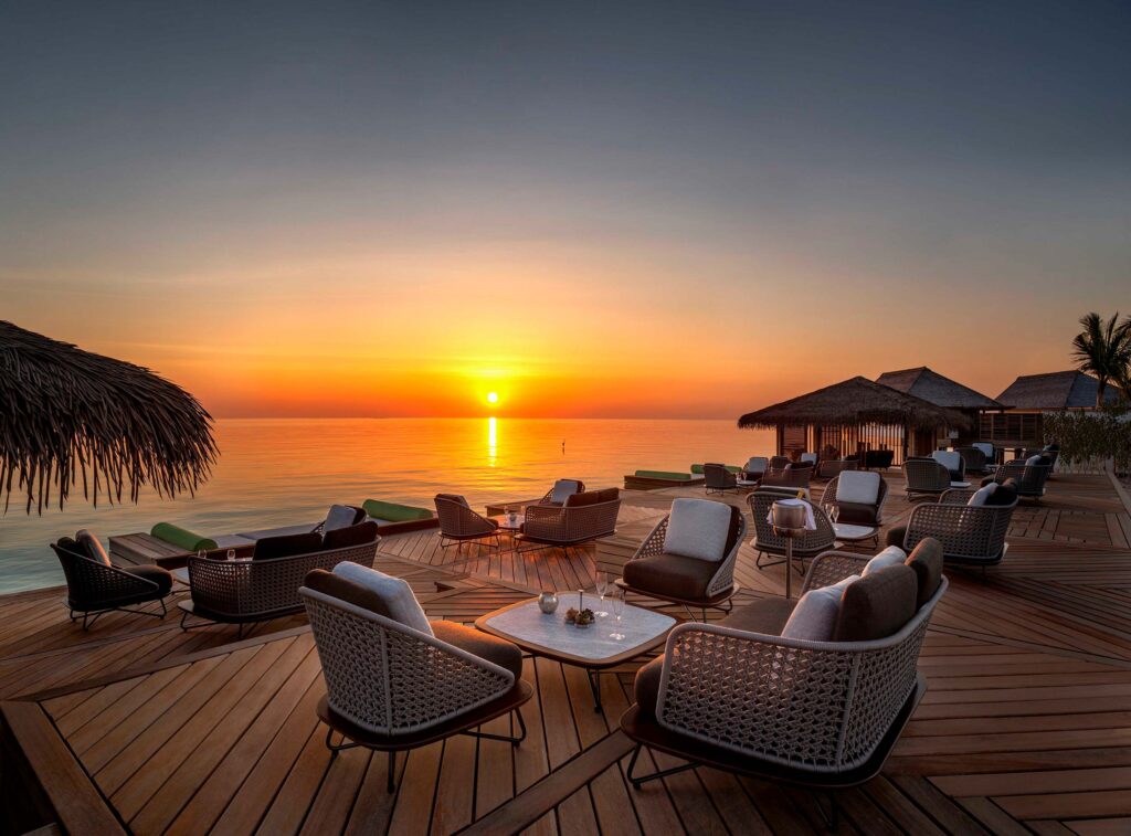 Terrasse am Wasser mit geflochtenen Sitzmöbeln und Holzboden vor Meer und orangefarbenem Sonnenuntergang