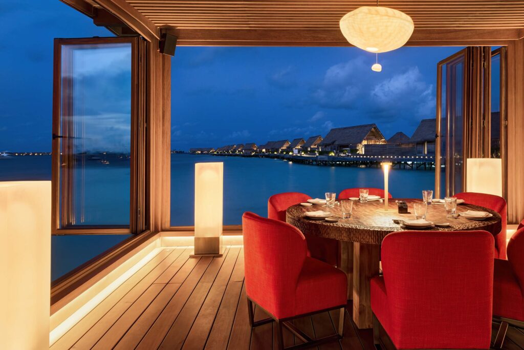 Hotelrestaurant mit rundem Holztisch, roten Stühlen, Holzplankenboden und Meerblick mit Wasserbungalows bei Abenddämmerung