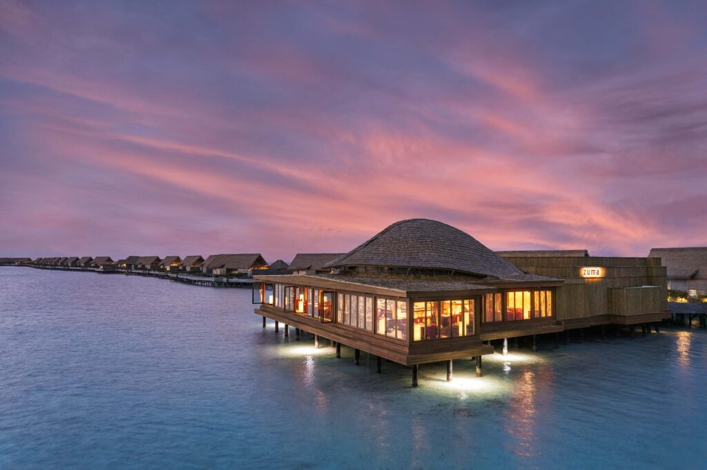 Reihe von Bungalows auf Stelzen über klarem Wasser mit beleuchtetem Holzgebäude und rosa-violettem Himmel