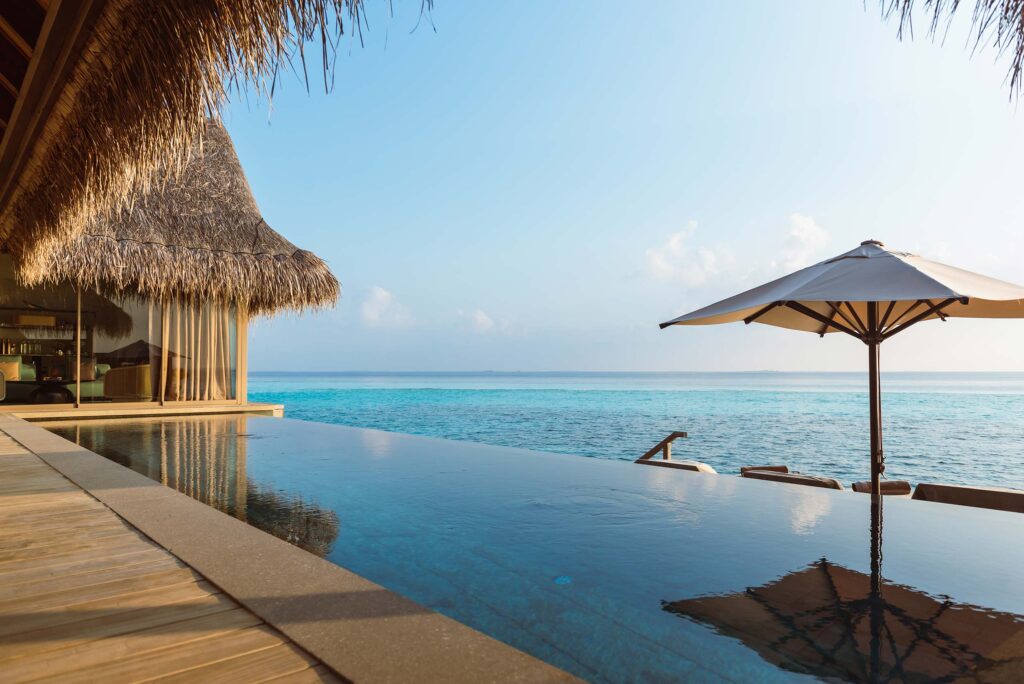 Infinity-Pool mit Holzdeck und strohgedecktem Gebäude links, großer Sonnenschirm und Meer im Hintergrund