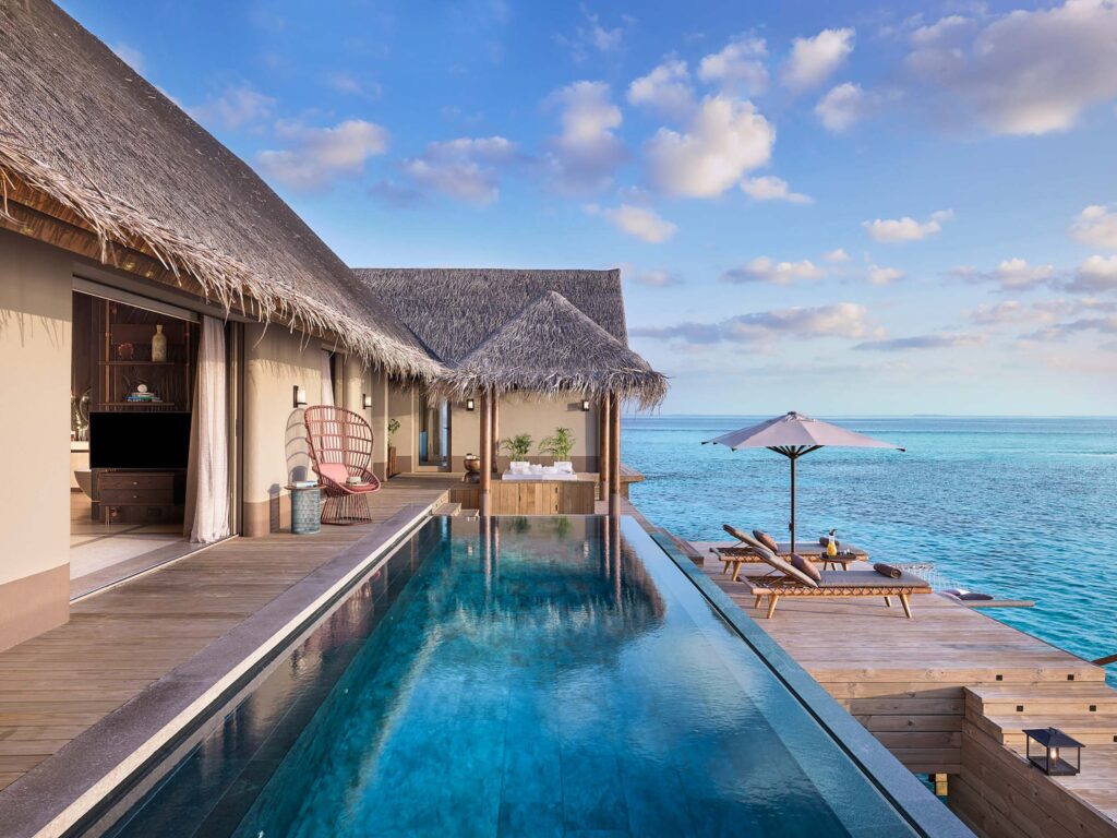 Infinity-Pool mit kristallklarem Wasser vor strohgedecktem Bungalow, Holzterrasse und Meer im Hintergrund