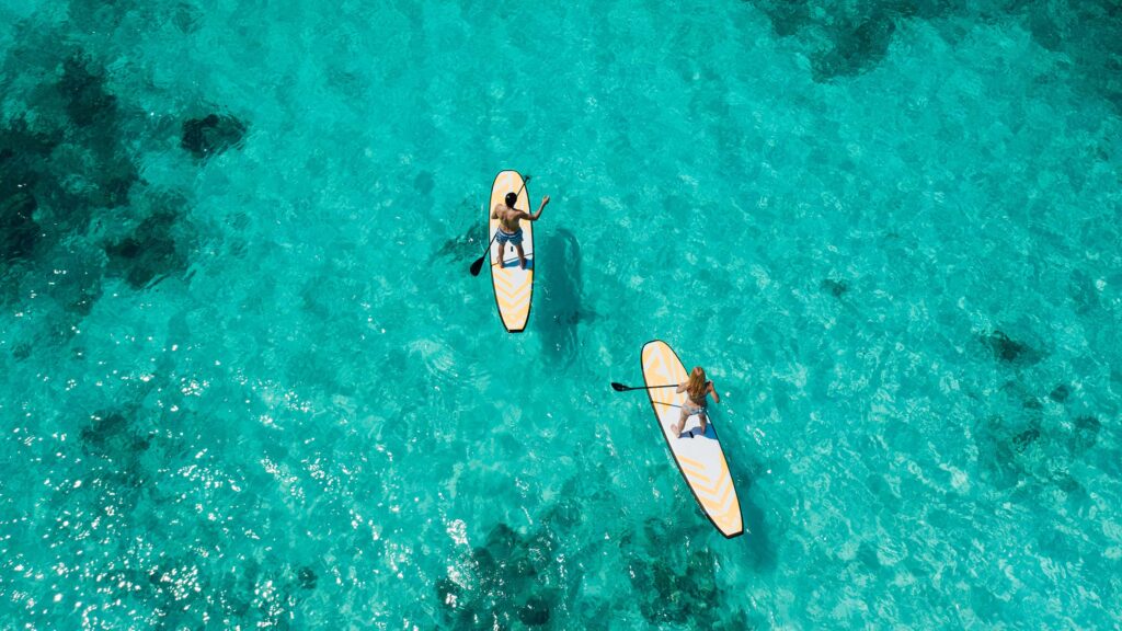 Zwei Personen auf weißen Stand-Up-Paddleboards mit orangenen Mustern gleiten über türkisblaues Wasser bei kräftigem.