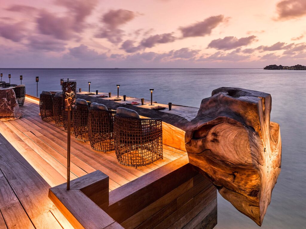 Terrasse mit Holztischen und Korbstühlen direkt am Wasser unter rosafarbenem Abendhimmel