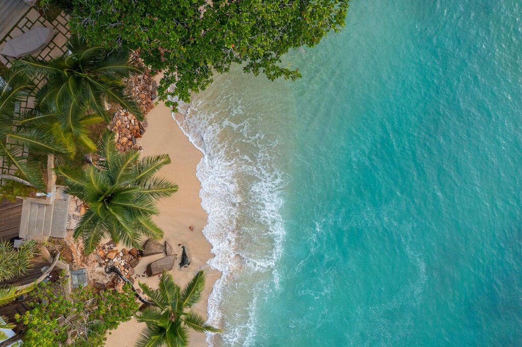 Luftaufnahme eines Strandes mit hellem Sand, türkisblauem Meer und Palmen an der Küste