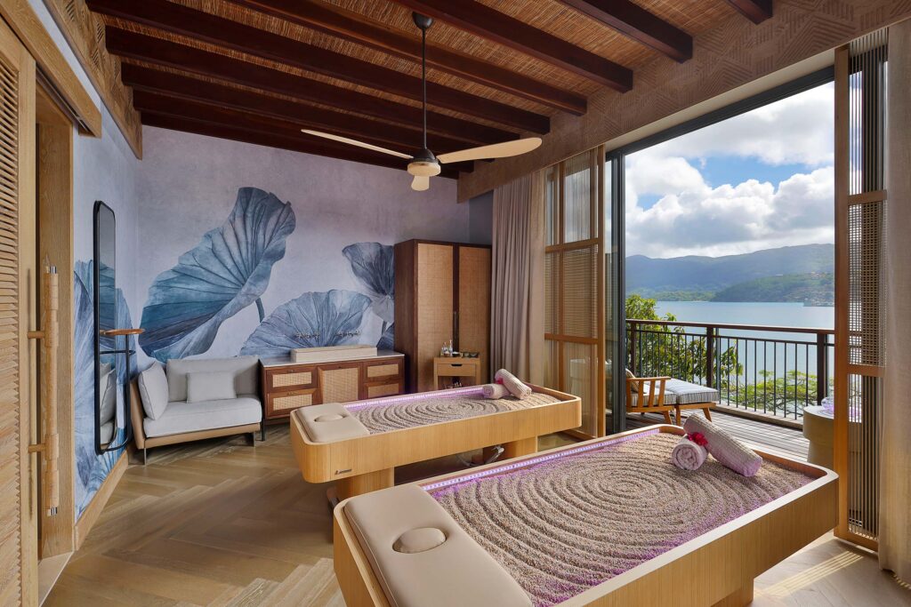 Heller Massageraum mit zwei Liegen, Holzfußboden, blauen Wandmotiven und Glasfront mit Wasserblick