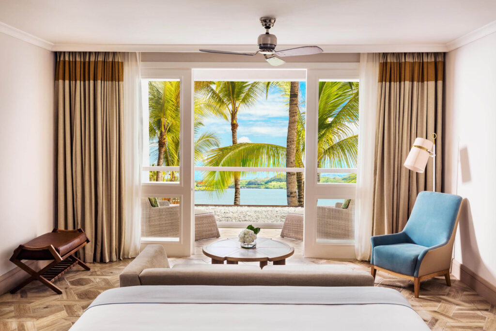 Hotelzimmer mit Bett, beige Couch, hellblauem Sessel, Stehlampe und Blick auf Palmen und Meer
