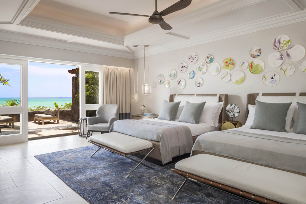 Geräumiges Hotelzimmer mit zwei Betten, dekorativen Tellern, blauem Teppich und großer Glastür zum Meer bei Tageslicht