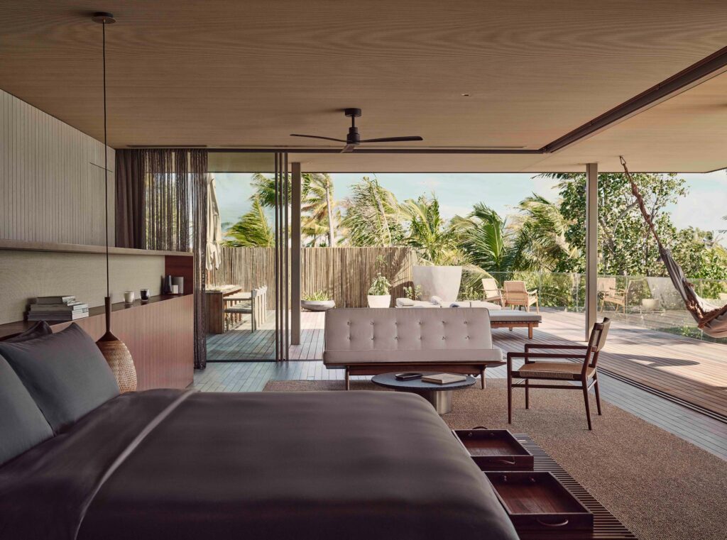 Modernes Hotelzimmer mit großem Bett, heller Couch, Holzsessel und großer Terrasse mit Blick auf grüne Palmen