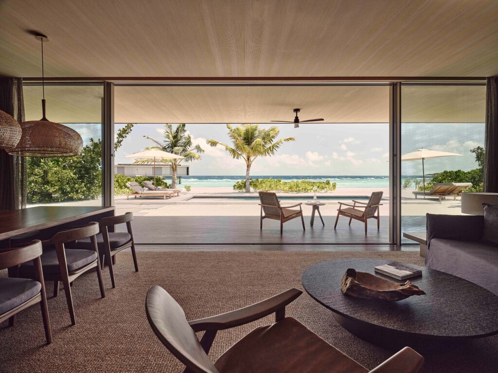 Modernes Hotelzimmer mit Holzmöbeln, großer Glasfront und Blick auf Terrasse mit Palmen und Meer