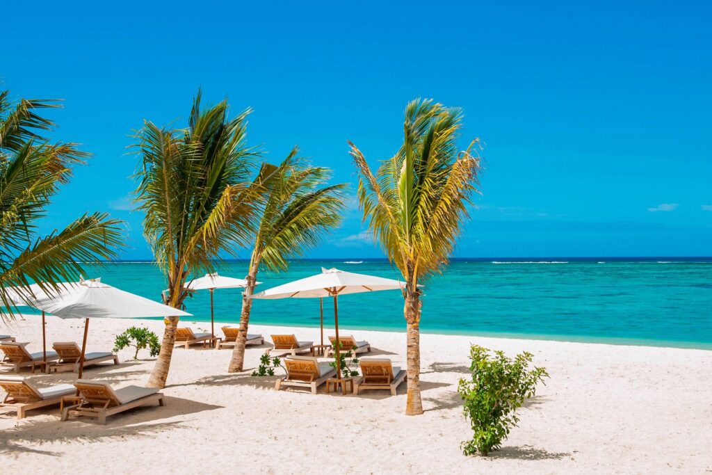 Sandstrand mit Holzliegen und weißen Stoffsonnenschirmen, Palmen im Vordergrund, türkisfarbenes Meer und blauer Himmel
