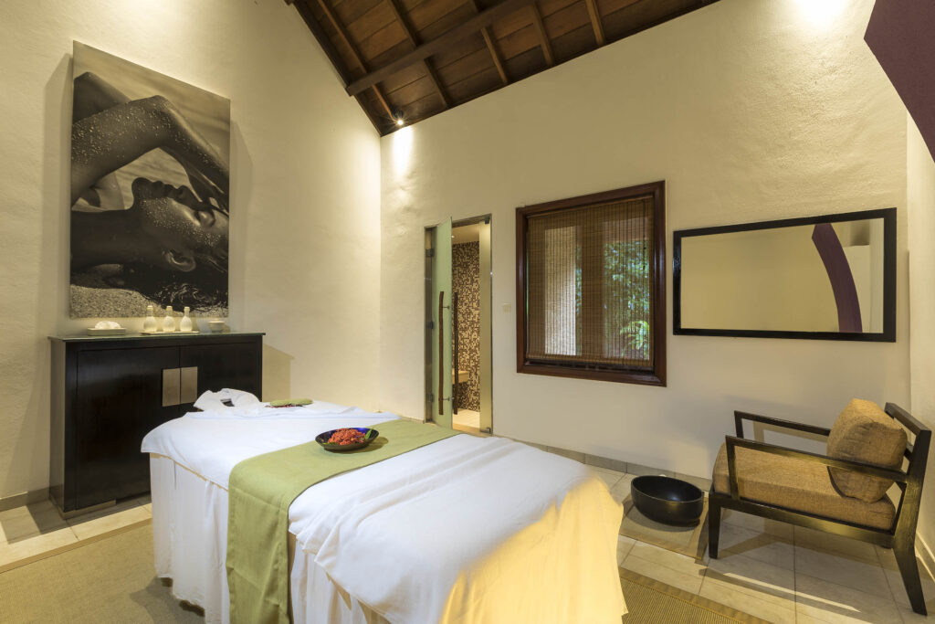 Massagezimmer mit Massagetisch, hölzernem Schrank, Sessel und warmem Licht im Shanti Maurice Resort & Spa