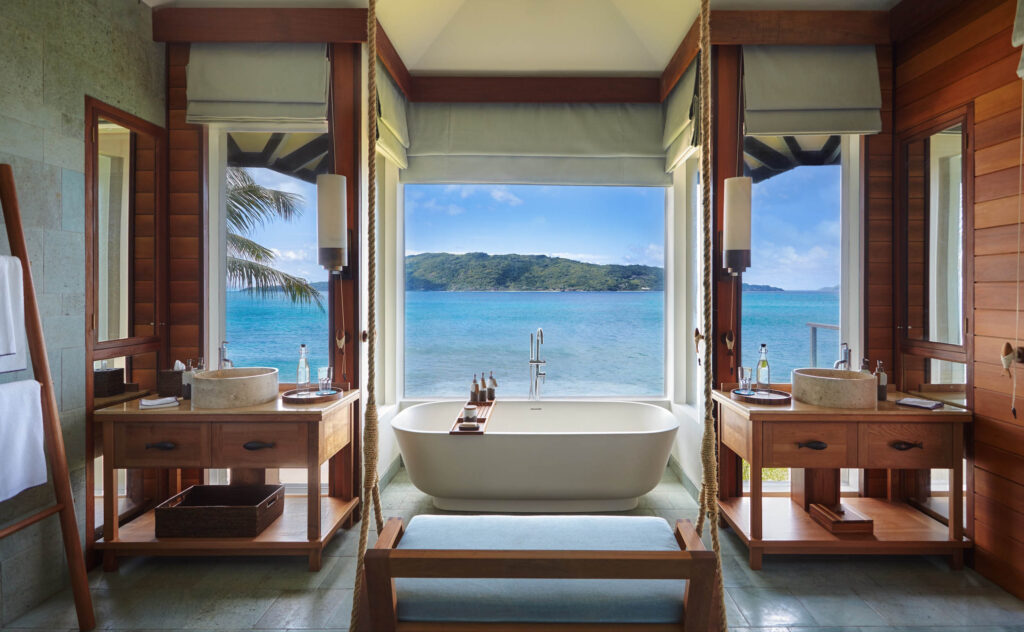 Badezimmer mit freistehender weißer Badewanne, Holzdecken und Steinboden mit Meerblick durch großes Fenster