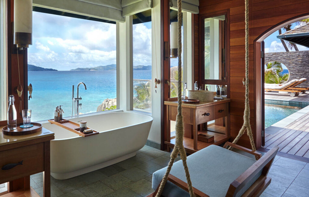 Badezimmer mit freistehender Badewanne, großen Fenstern, Holztisch und Blick auf türkisfarbenes Meer