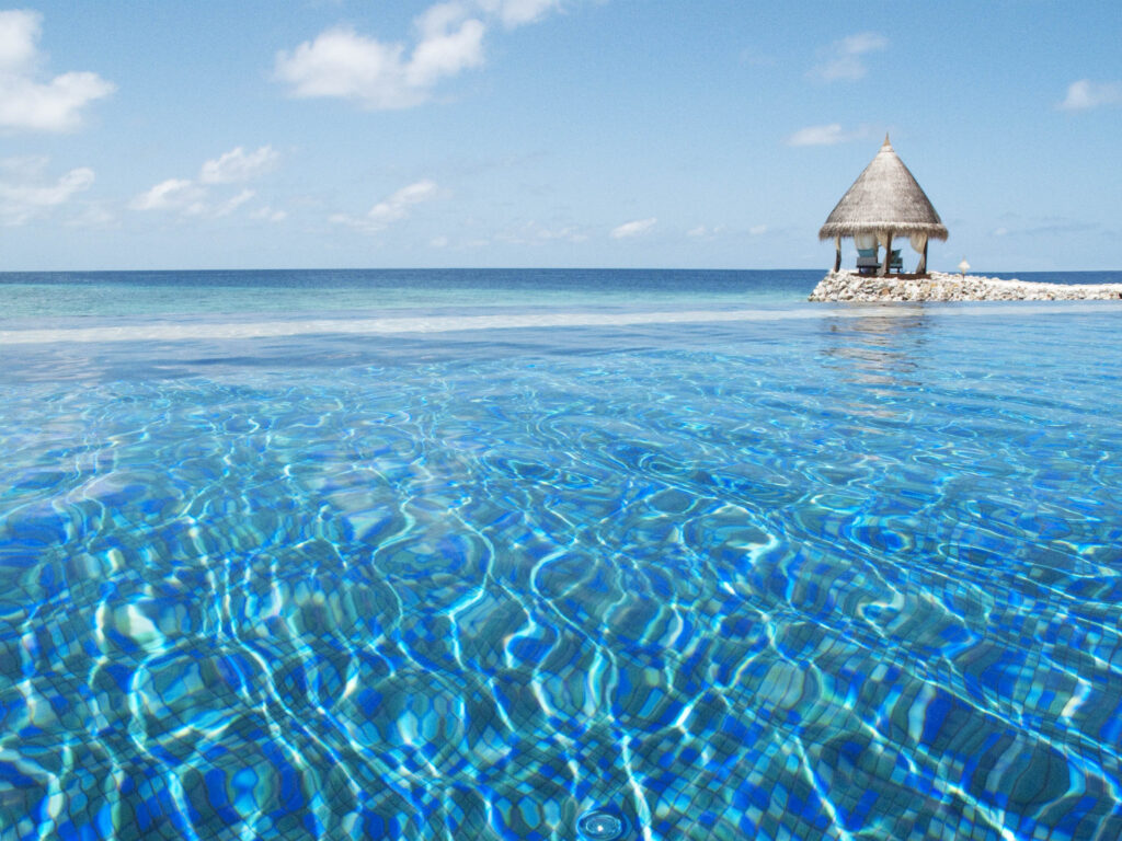 Infinity-Pool mit blauem Wasser und Lichtreflexionen, kleiner strohgedeckter Pavillon auf Steinplattform im Hintergrund