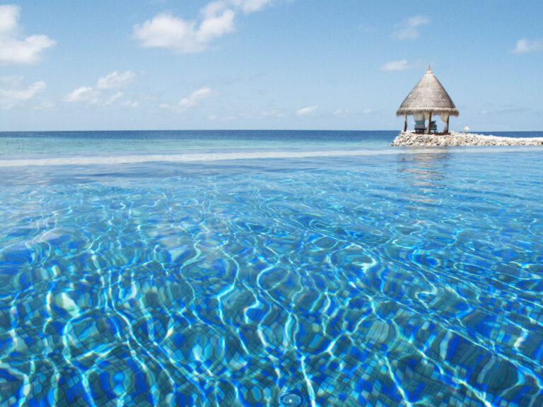Infinity-Pool mit blauem Wasser und Lichtreflexionen, kleiner strohgedeckter Pavillon auf Steinplattform im Hintergrund
