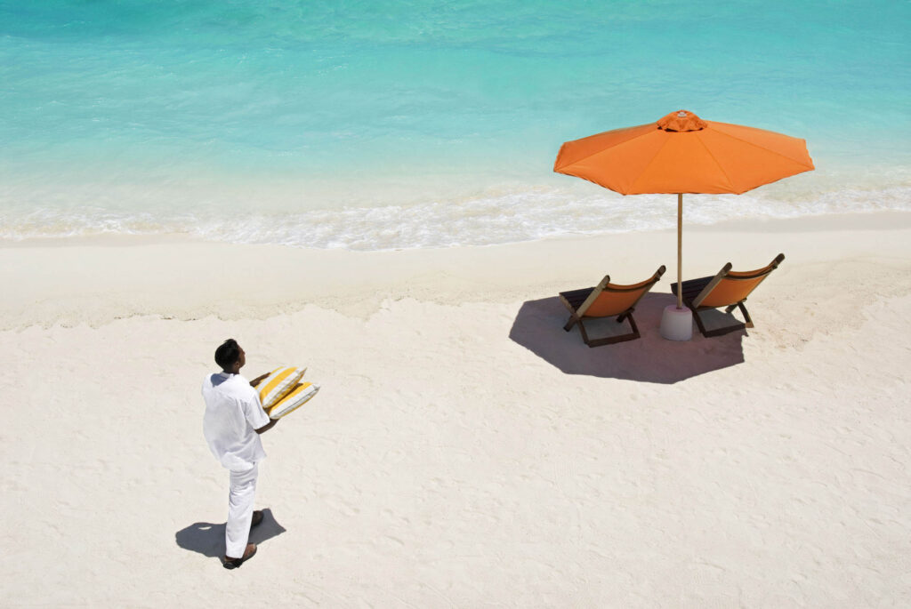 Strand mit weißem Sand, zwei hölzernen Liegestühlen mit orangefarbenem Stoff unter orangem Sonnenschirm am türkisblauen Meer