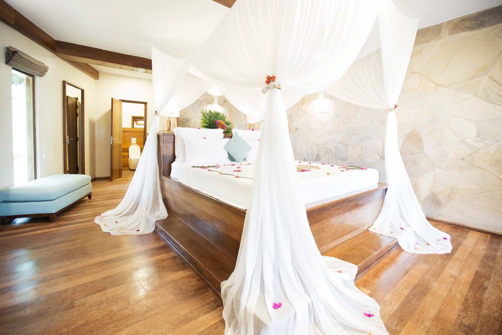 Hell beleuchtetes Hotelzimmer mit großem Holz-Himmelbett, weißen Vorhängen und verstreuten Blumenblüten