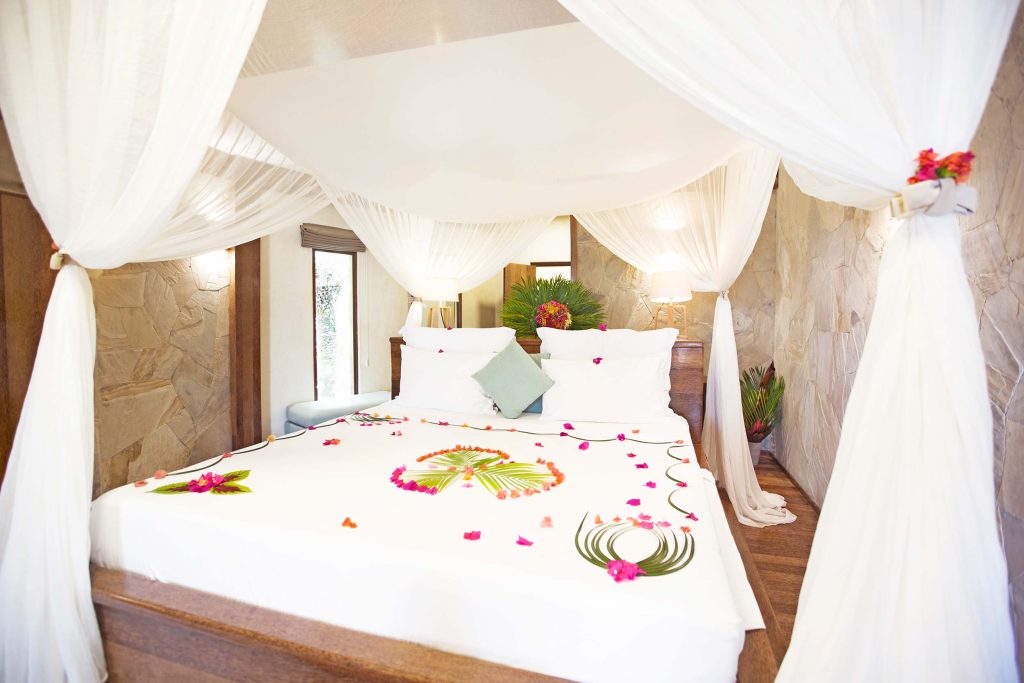 Hotelzimmer in Tulia Zanzibar mit Himmelbett, weißen Vorhängen, Natursteinwänden und dekorativen Blumenarrangements